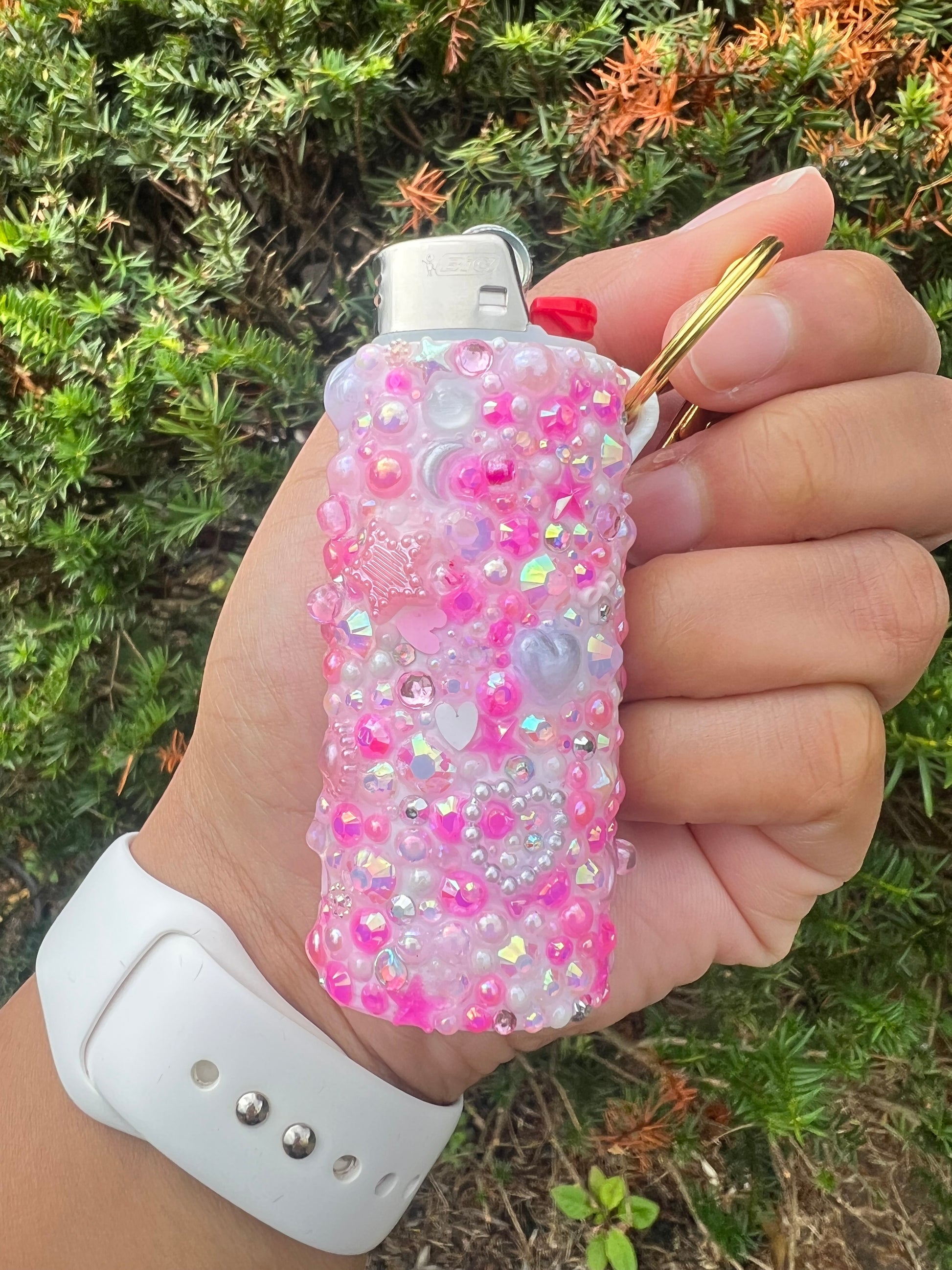 Hand holding a Bic lighter in a clear junk charm case covered in pink rhinestones pearls and iridescent gems with a gold keychain ring