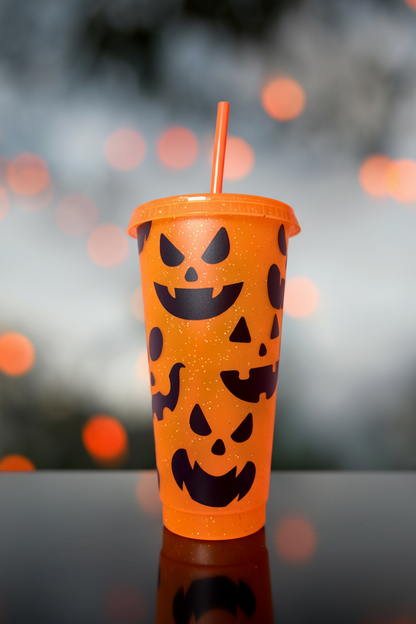 Orange tumbler with black jack-o'-lantern faces on a reflective surface with blurred lights in the background.