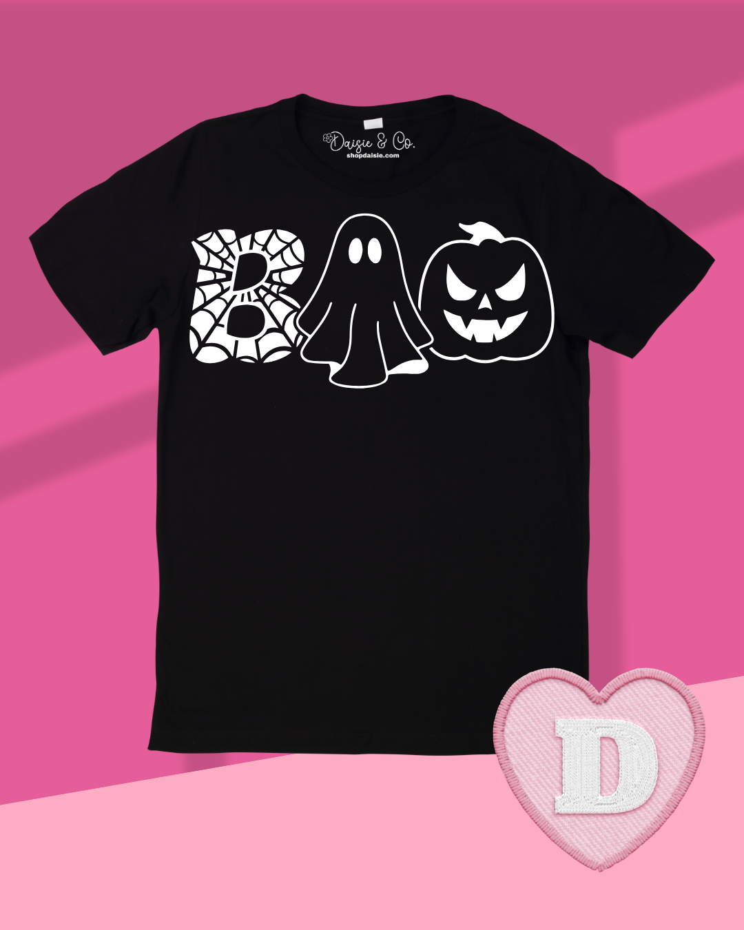 A black short-sleeve t-shirt with a white graphic spelling 'BOO' using a spiderweb 'B', a ghost, and a jack-o'-lantern 'O'. The shirt is displayed flat against a pink background with a pink heart-shaped patch in the bottom right corner.