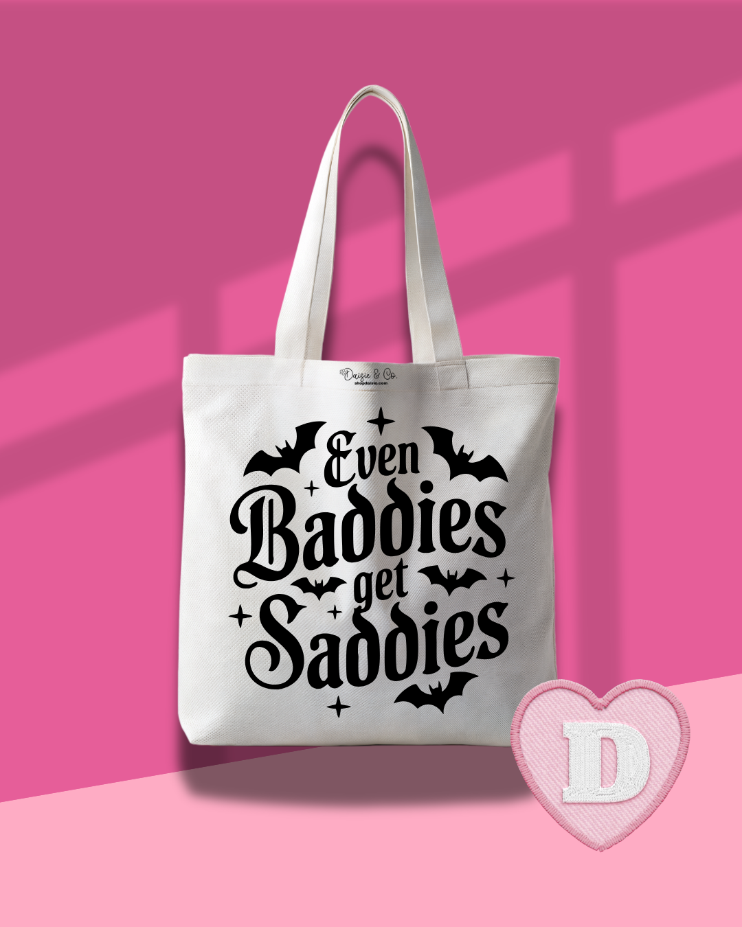 A white canvas tote bag with black gothic text reading 'Even Baddies get Saddies' and small bat silhouettes, shown against a pink background with a pink heart-shaped watermark in the lower right.