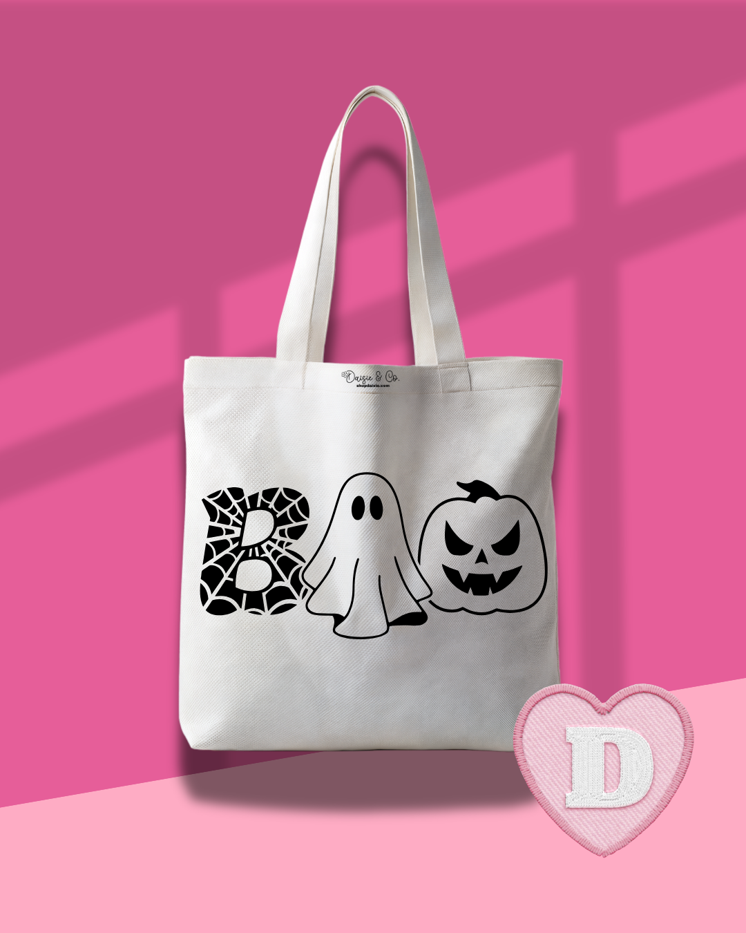 A white canvas tote bag with black Halloween-themed graphics spelling 'BOO' using a spiderweb, a ghost, and a jack-o'-lantern. The bag is displayed against a pink background with window shadow overlays and a pink heart-shaped logo overlay in the bottom right corner.