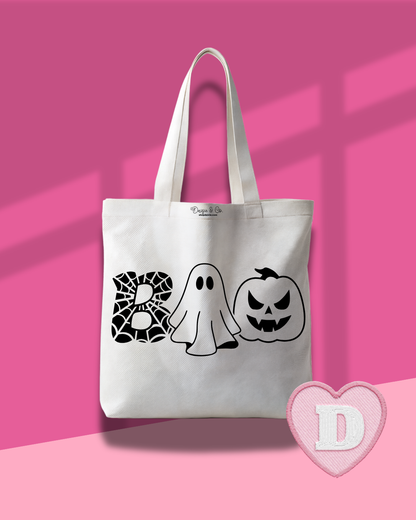 A white canvas tote bag with black Halloween-themed graphics spelling 'BOO' using a spiderweb, a ghost, and a jack-o'-lantern. The bag is displayed against a pink background with window shadow overlays and a pink heart-shaped logo overlay in the bottom right corner.