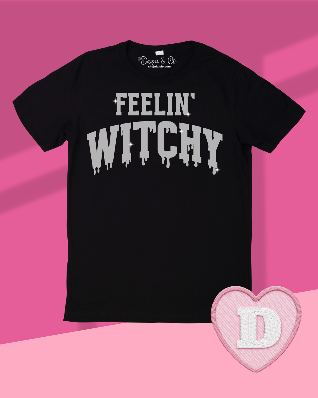 A black short-sleeve t-shirt with 'FEELIN' WITCHY' printed in a sparkly silver dripping font across the chest, displayed on a pink and light pink background with a pink heart-shaped patch featuring a white letter 'D' in the lower right corner.