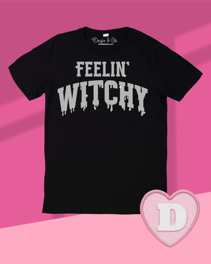 A black short-sleeve t-shirt with 'FEELIN' WITCHY' printed in a sparkly silver dripping font across the chest, displayed on a pink and light pink background with a pink heart-shaped patch featuring a white letter 'D' in the lower right corner.