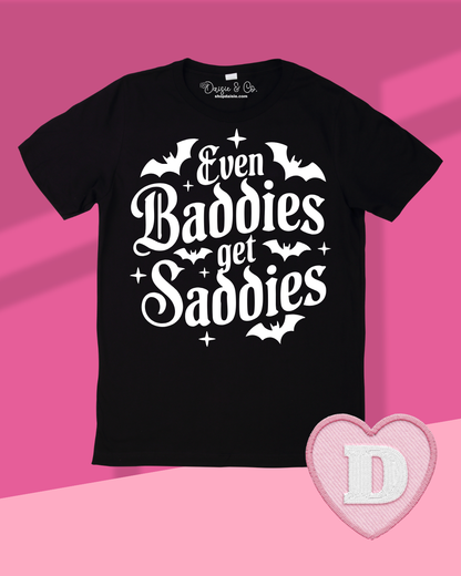 A black short-sleeve graphic T-shirt laid flat on a pink background, featuring the phrase 'Even Baddies get Sadies' in large white decorative lettering with bat and star accents. A pink heart-shaped patch with a white letter D appears in the lower right corner.