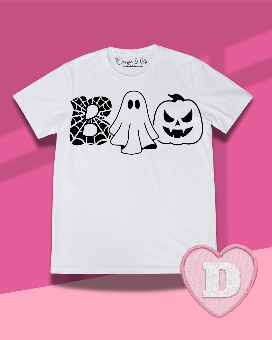 A white short-sleeve t-shirt with a black graphic spelling 'BOO' using a spiderweb, a ghost, and a jack-o'-lantern. The shirt is displayed flat against a pink background with a pink heart-shaped patch featuring the letter 'D' in the bottom right corner.