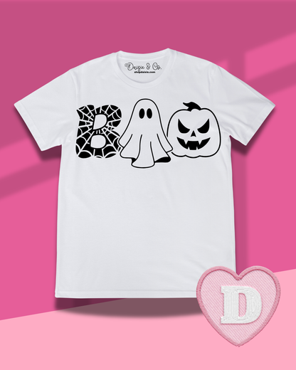 A white short-sleeve t-shirt with a black graphic spelling 'BOO' using a spiderweb, a ghost, and a jack-o'-lantern. The shirt is displayed flat against a pink background with a pink heart-shaped patch featuring the letter 'D' in the bottom right corner.