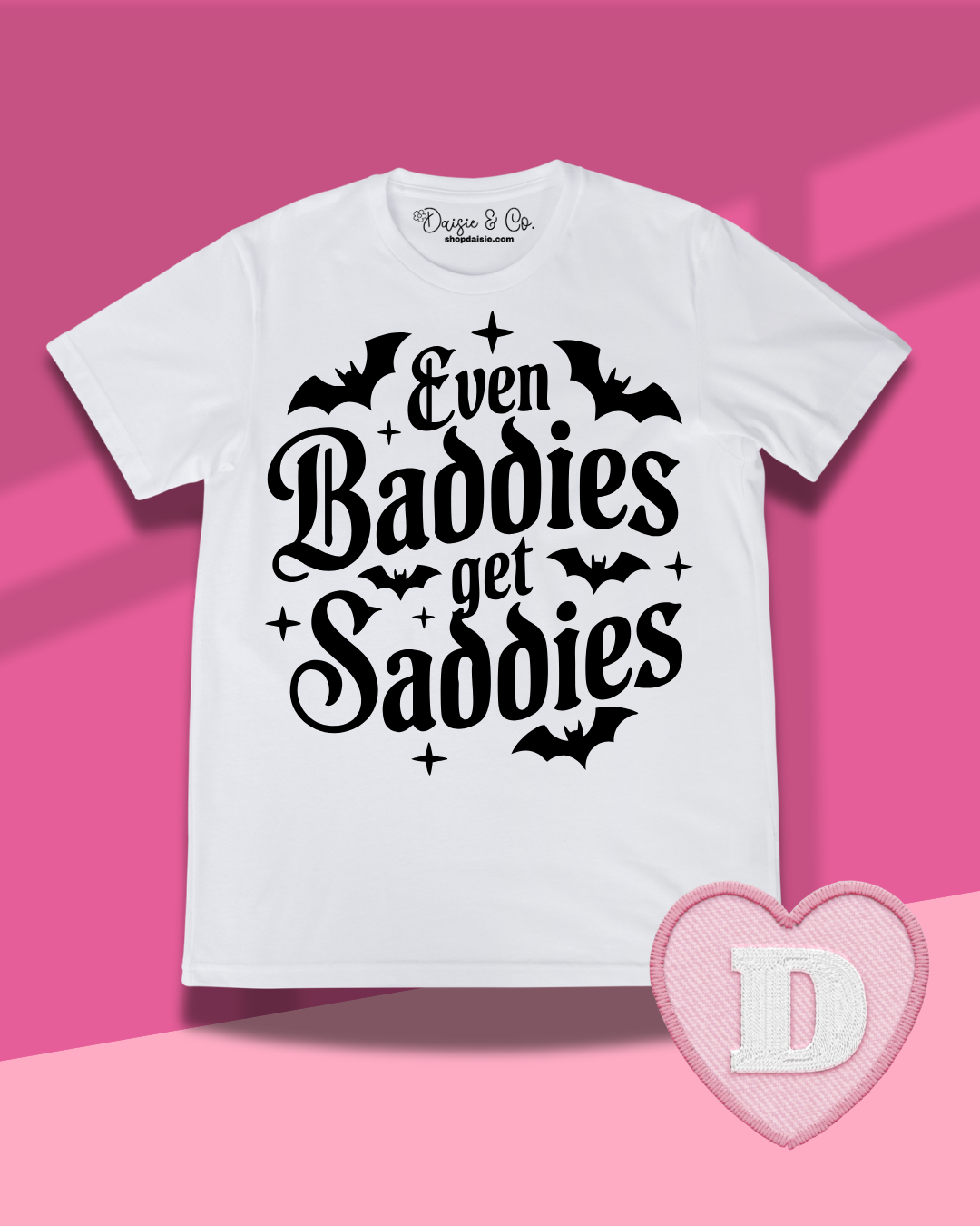 A white short-sleeve graphic T-shirt with black text reading 'Even Baddies get Saddies' surrounded by bat silhouettes and small star accents, shown on a pink background. A pink heart-shaped patch with a white letter D appears in the lower right corner.